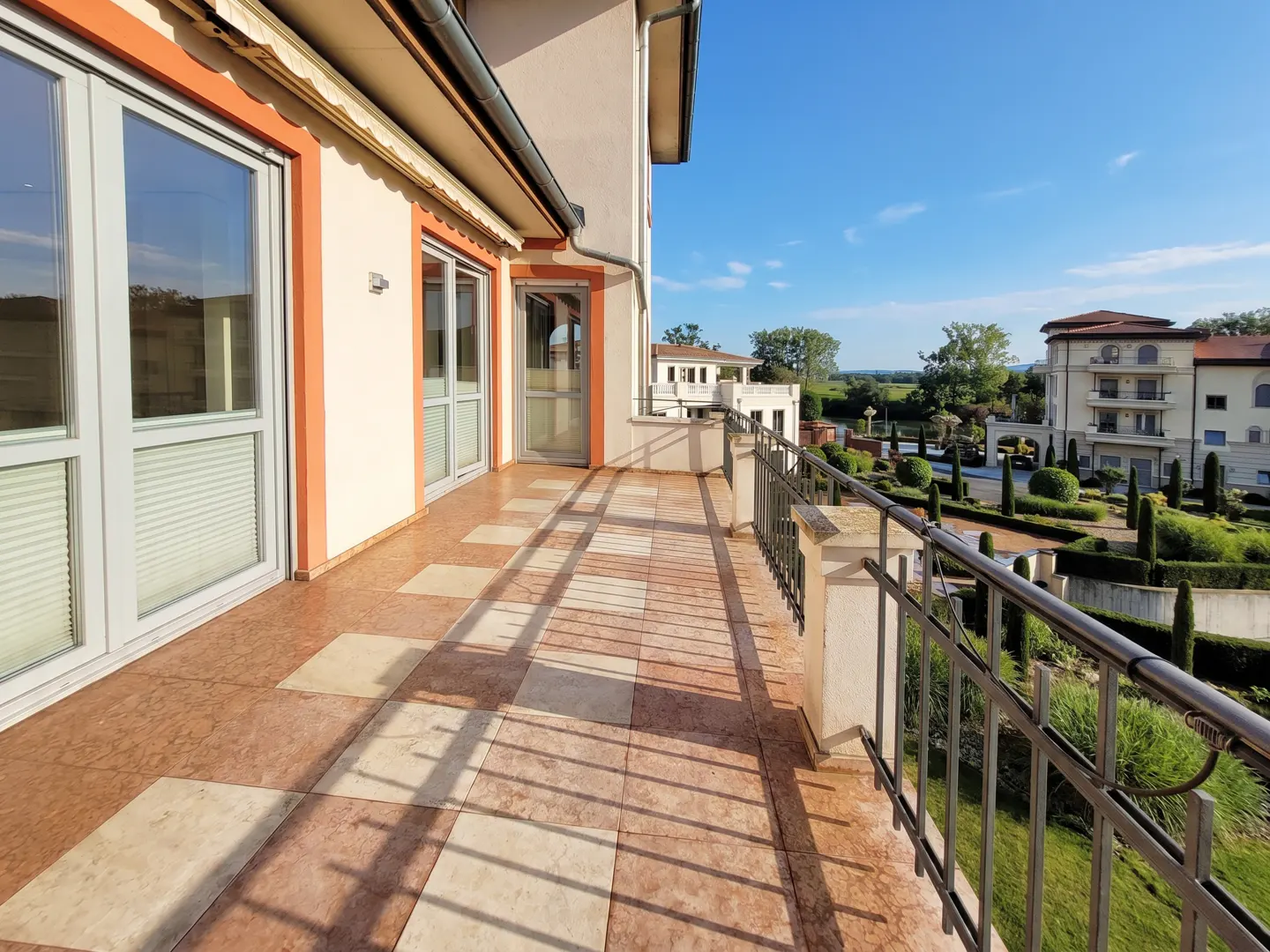 Balcony view with orange trim, tiled floor, and metal railing overlooking manicured gardens and buildings under a blue sky.