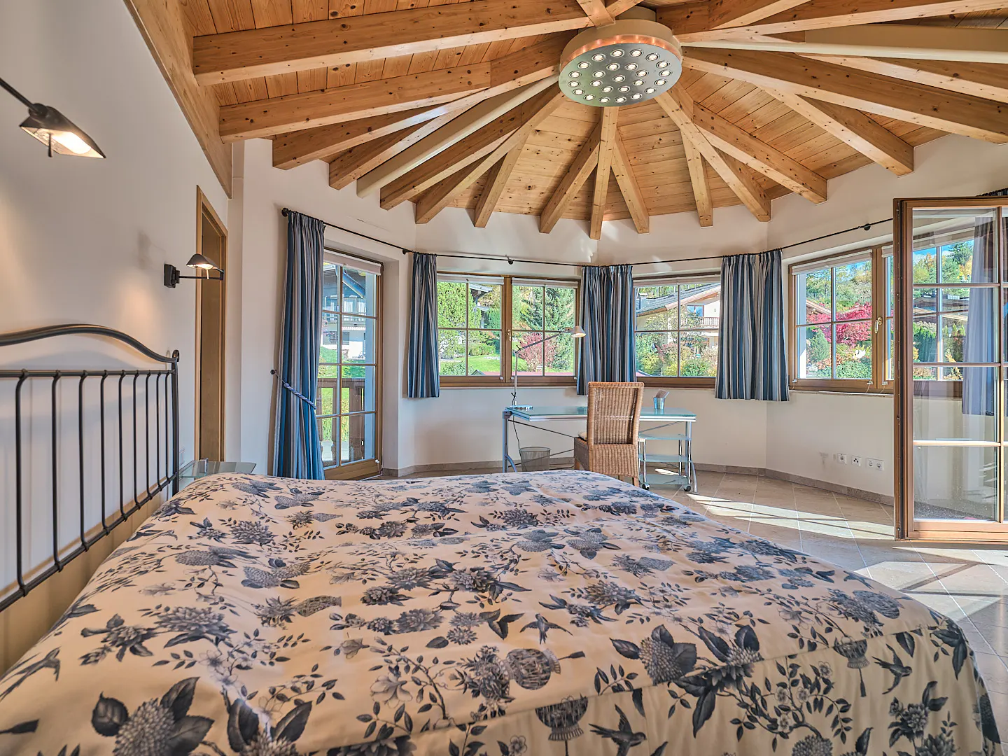 Bedroom with floral bedding, iron bed frame, wood ceiling, and windows with blue curtains overlooking a green yard.