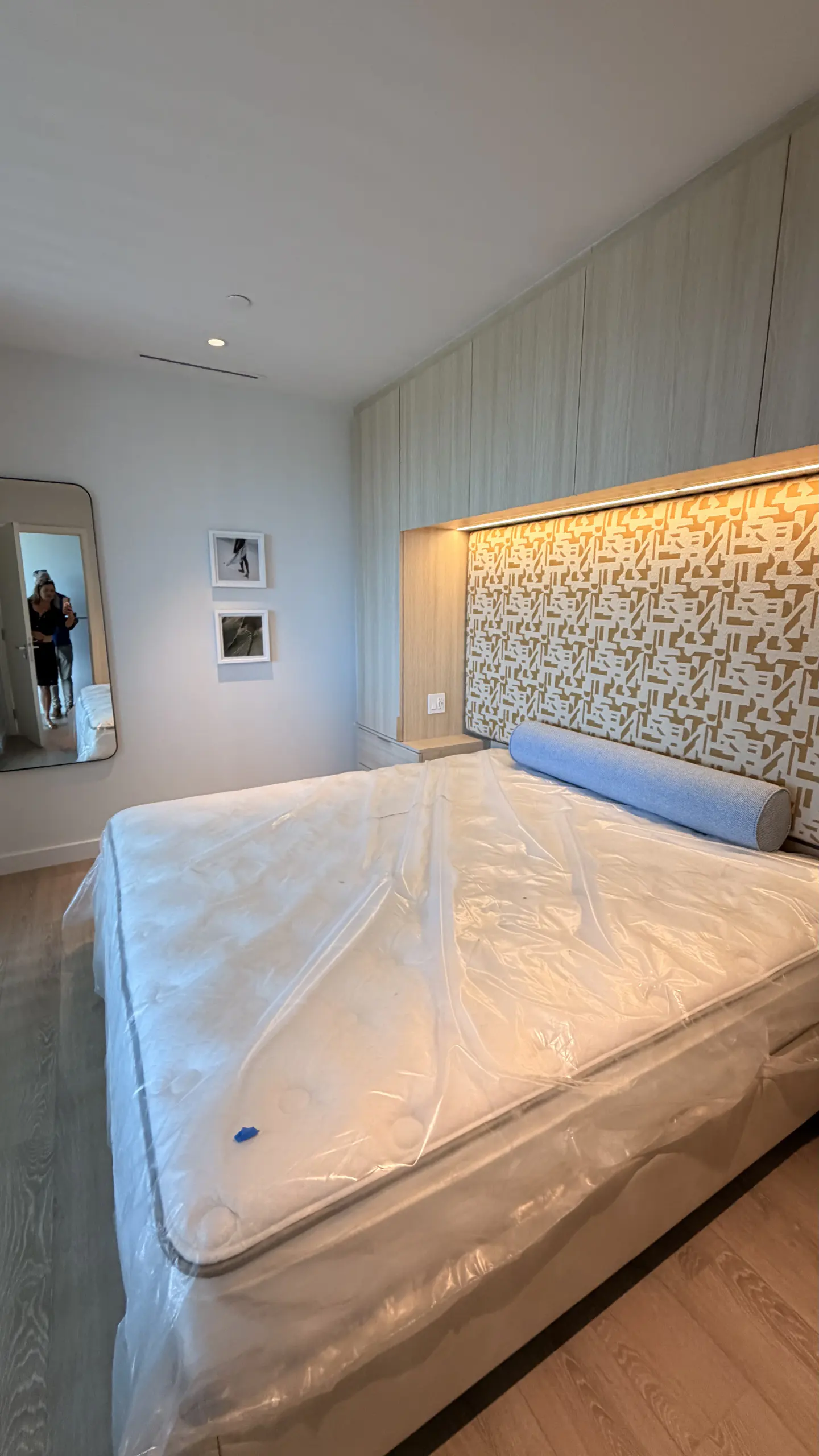 Bedroom with a large bed covered in plastic, a patterned headboard with lighting, and a mirror reflecting people.