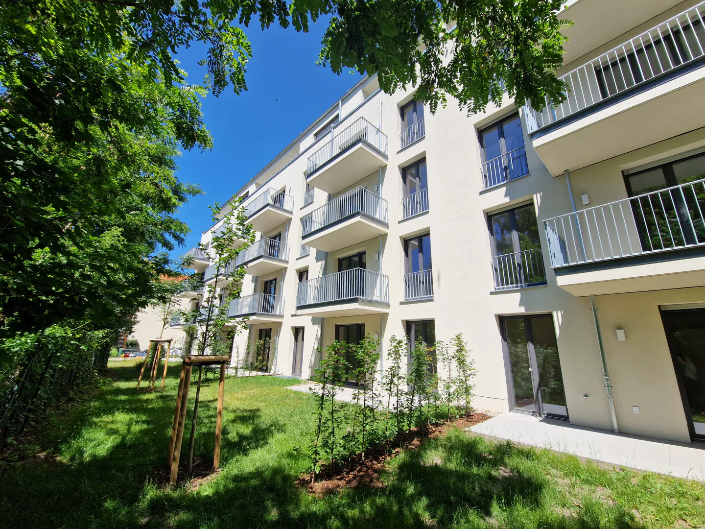 Modern apartment building with white facade and balconies, surrounded by green lawn and trees under a blue sky.