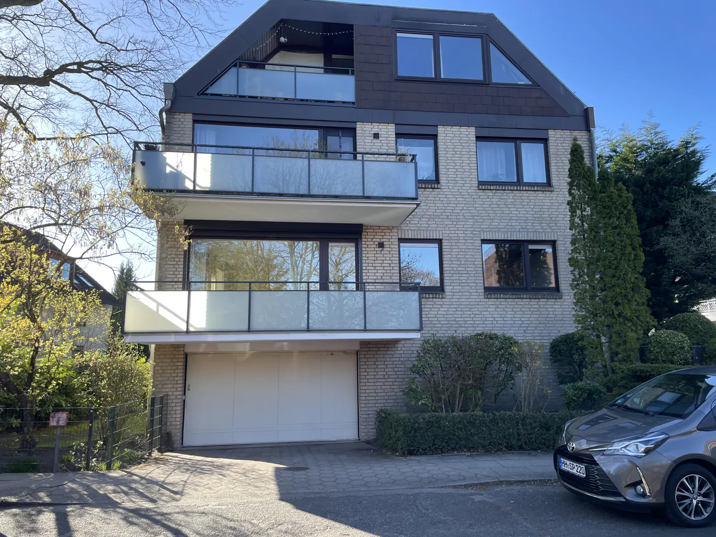 Three-story brick house with balconies and a garage, surrounded by greenery and a gray car parked nearby.