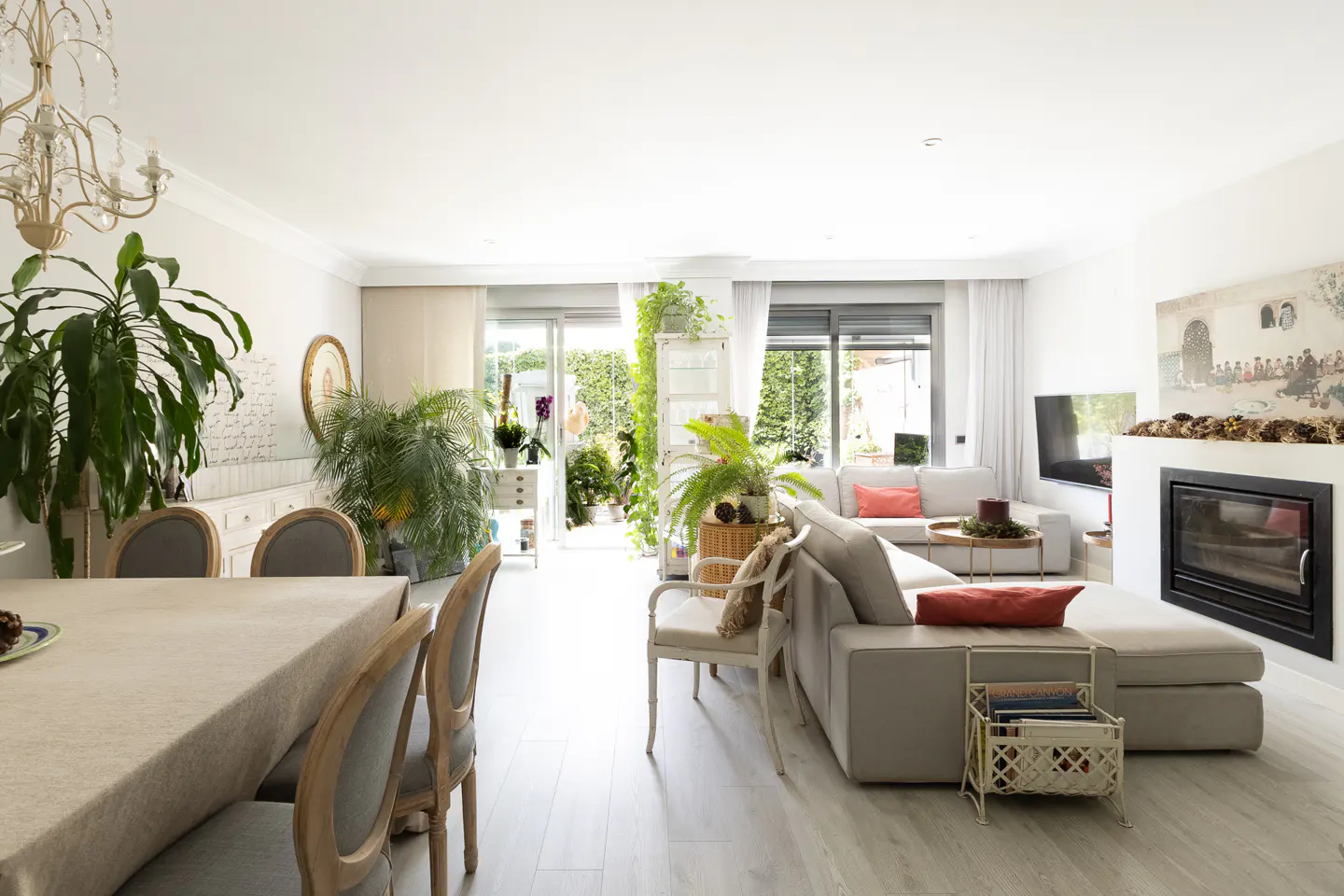 Bright living room with dining area, beige sofa, fireplace, and large windows leading to a lush green patio.