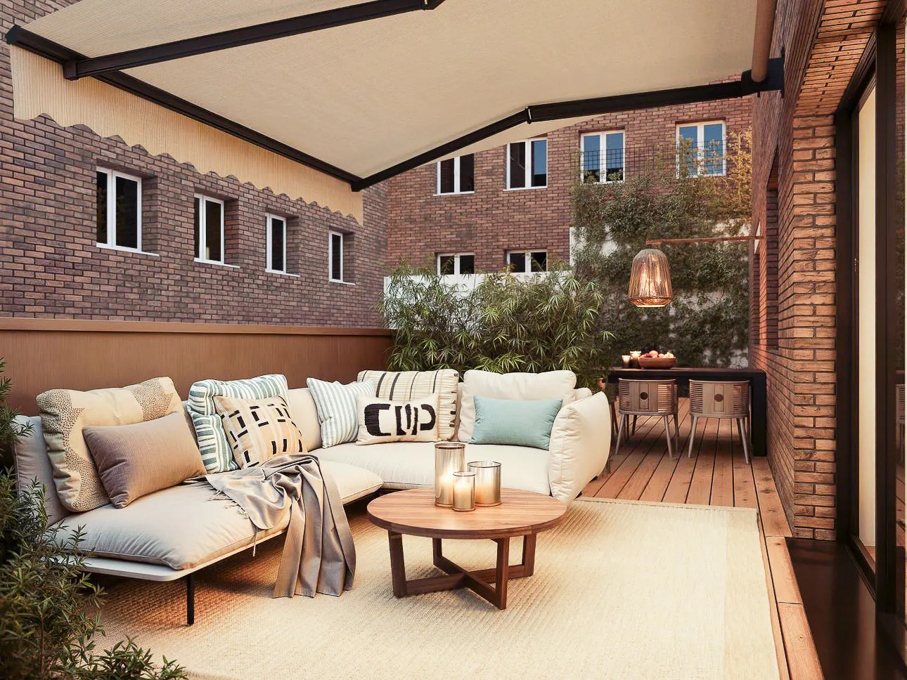 Outdoor patio with a cream sofa, pillows, and a round wooden table with candles. Brick buildings and greenery in the background.