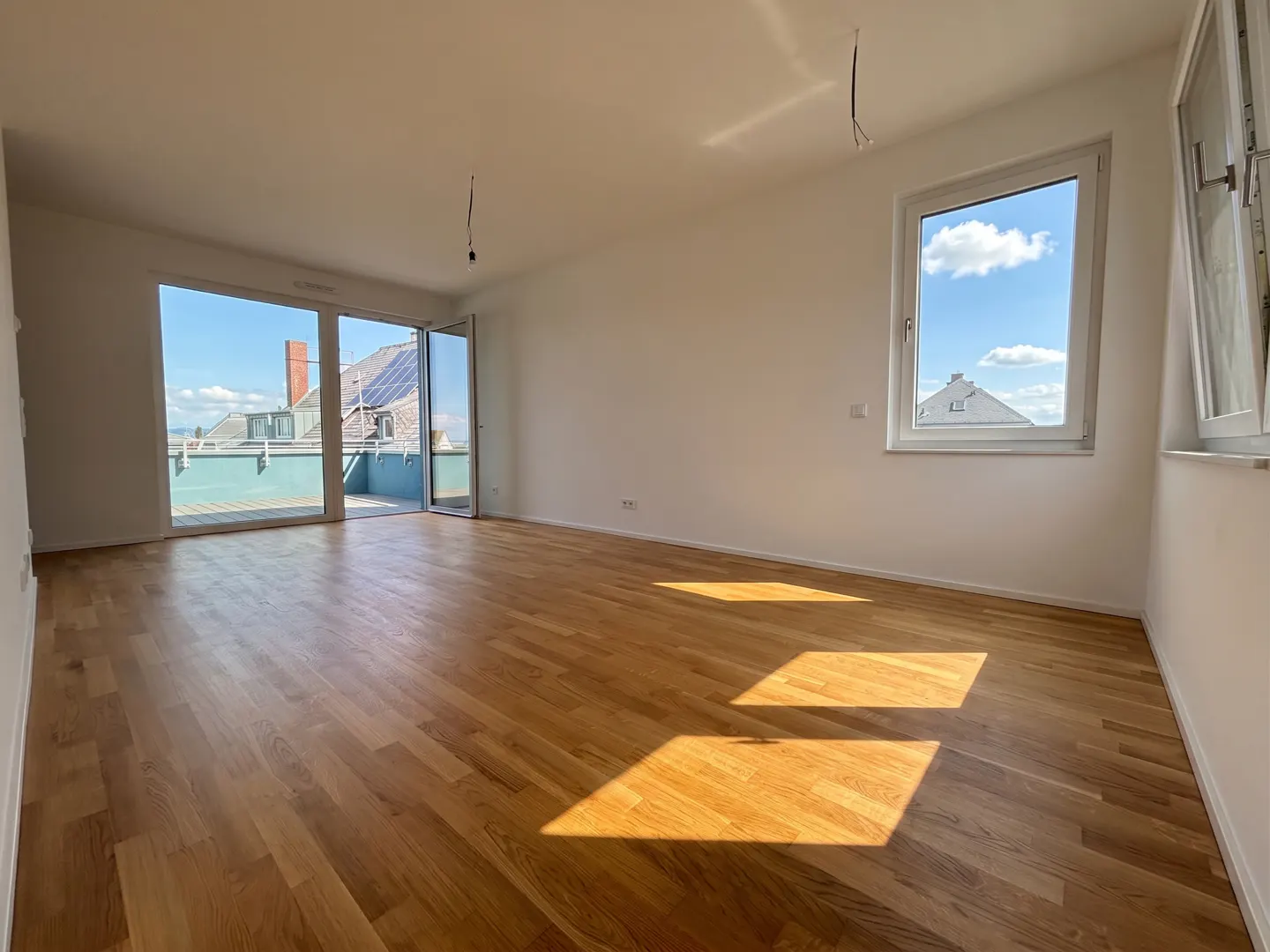 Bright, empty room with light wood floors, white walls, and a balcony with a view of rooftops and blue sky. Sunlight streams in.