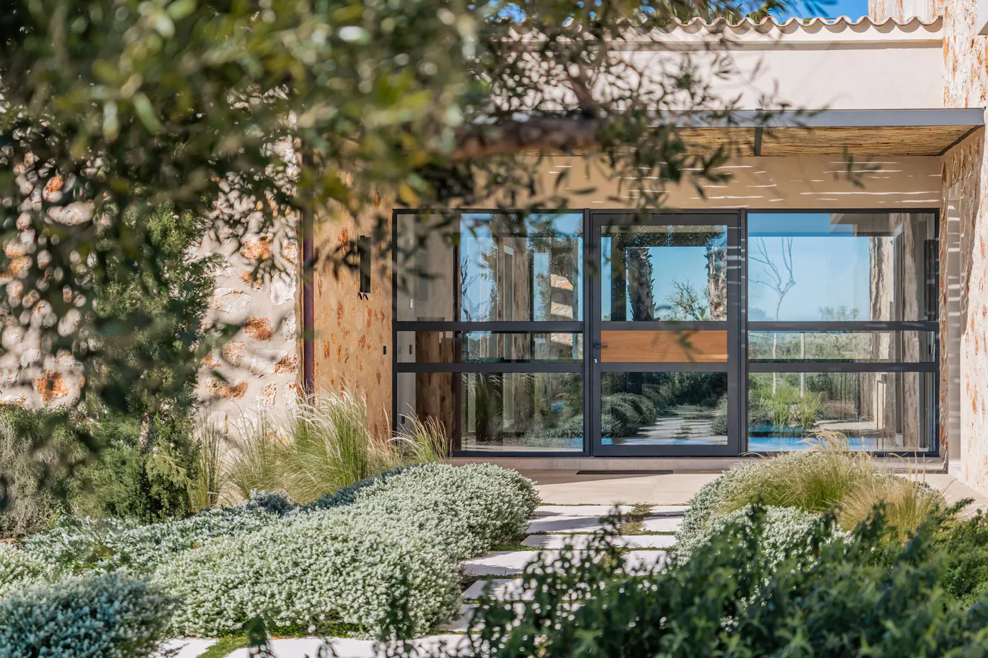 Modern home exterior with stone walls, a black framed glass door, and a stone path through lush green landscaping.
