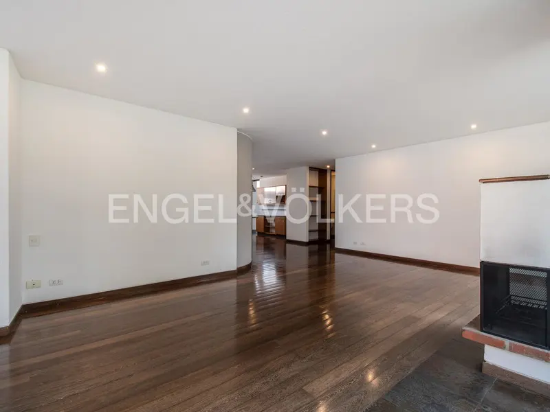 Bright, empty living room with hardwood floors, white walls, and recessed lighting. A fireplace is on the right, and a kitchen is visible in the background.