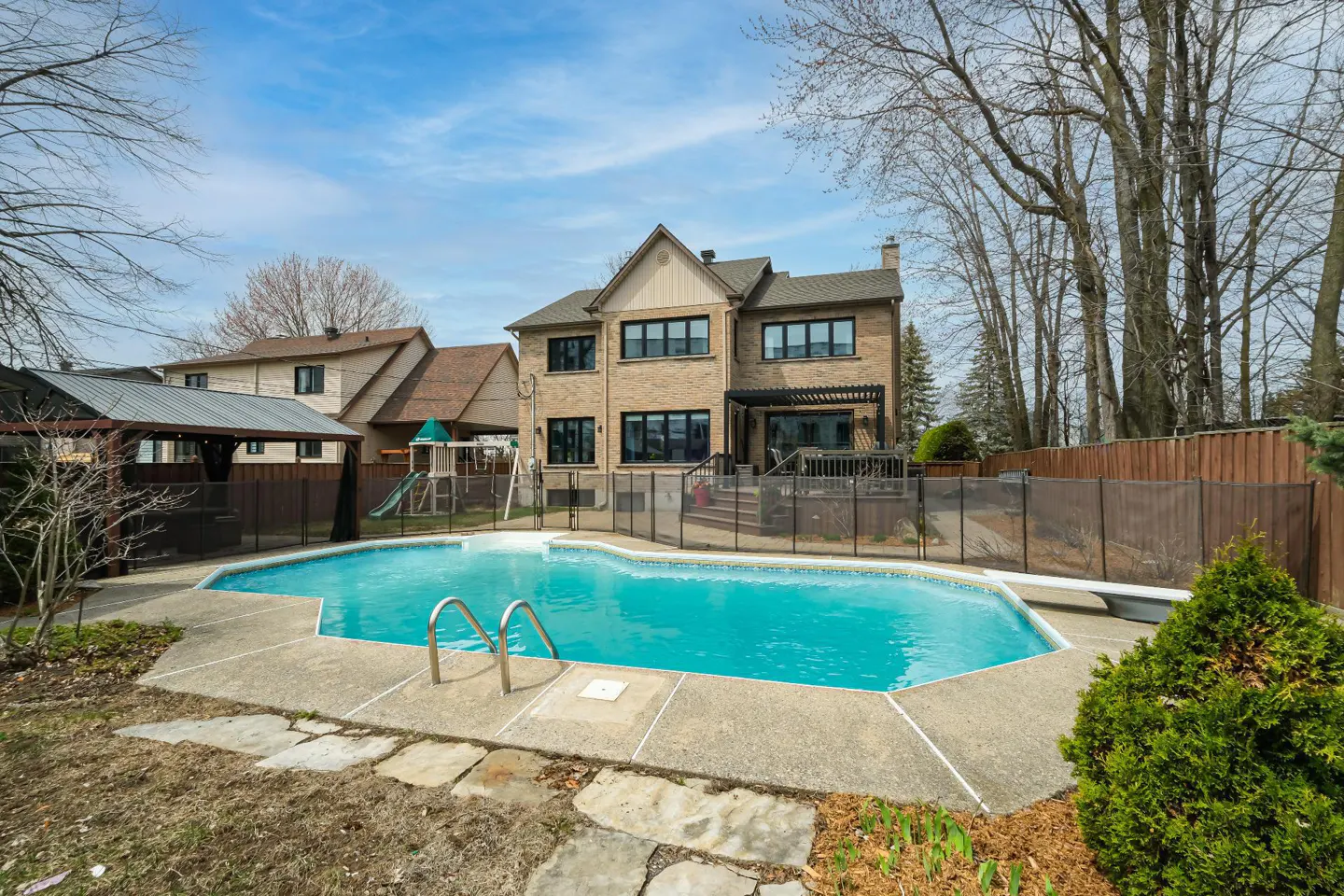 Backyard view of a two-story brick house with a blue swimming pool, surrounded by a fence, patio, and trees.