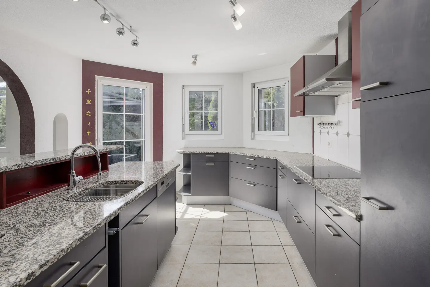 A modern kitchen with gray cabinets, granite countertops, and white tile flooring. Natural light streams in through multiple windows.