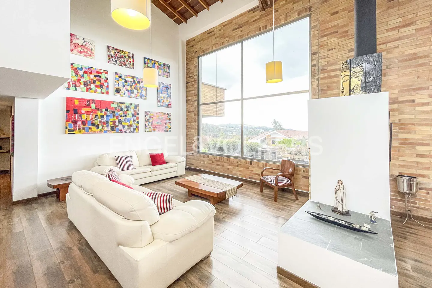 Bright living room with white sofas, wood floors, and brick wall with large window. Colorful art adorns the wall.