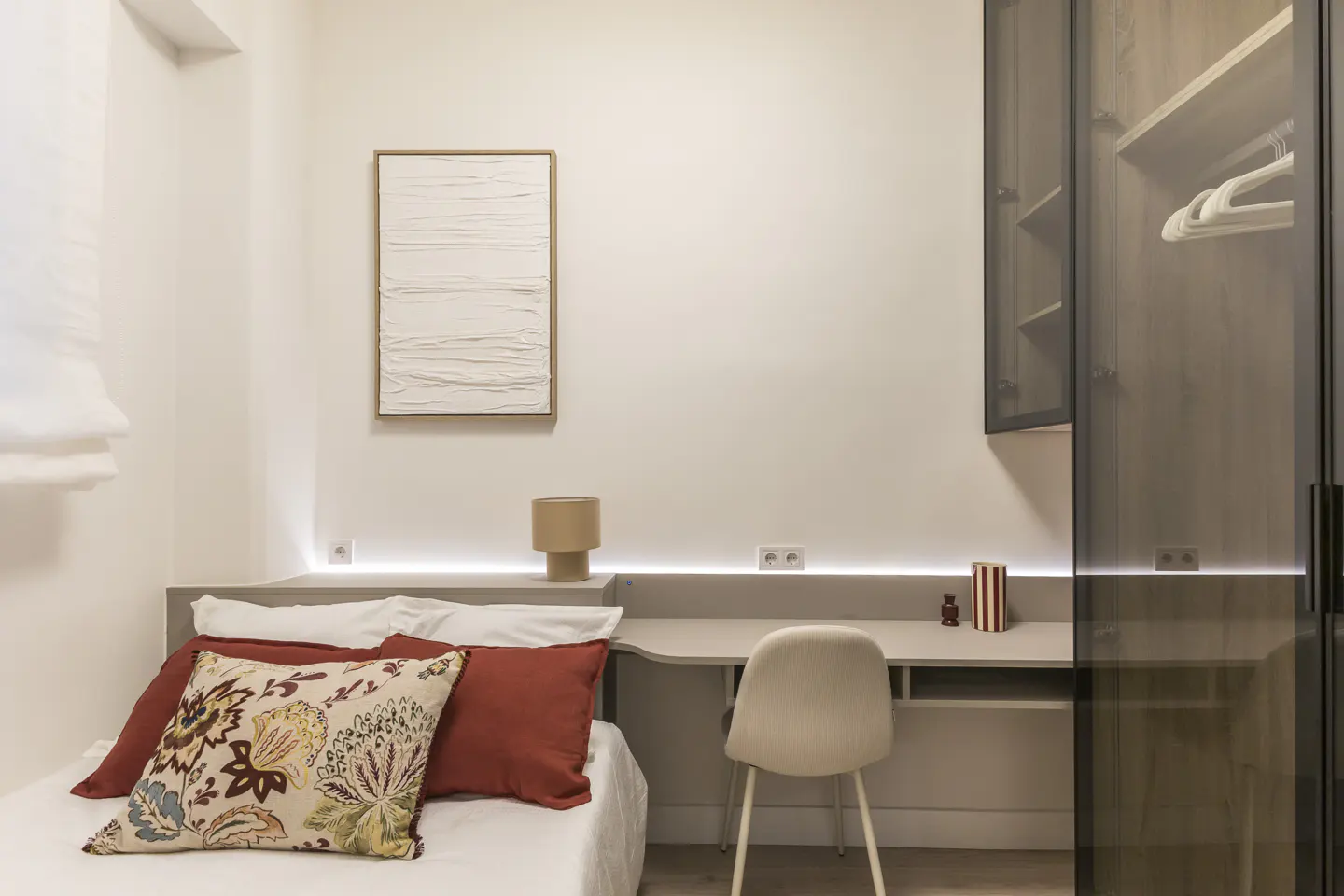 A bedroom with a white bed, red pillows, a desk, a chair, and a closet with smoked glass doors.