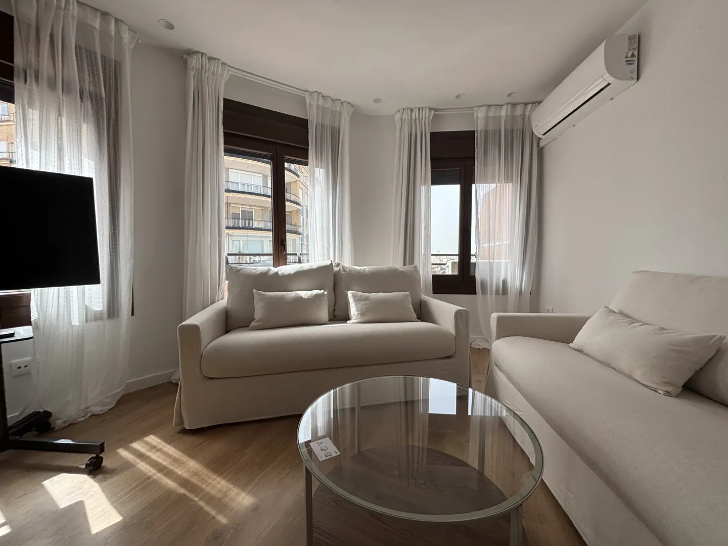 Bright living room with two cream sofas, a glass coffee table, and sheer white curtains over dark-framed windows.
