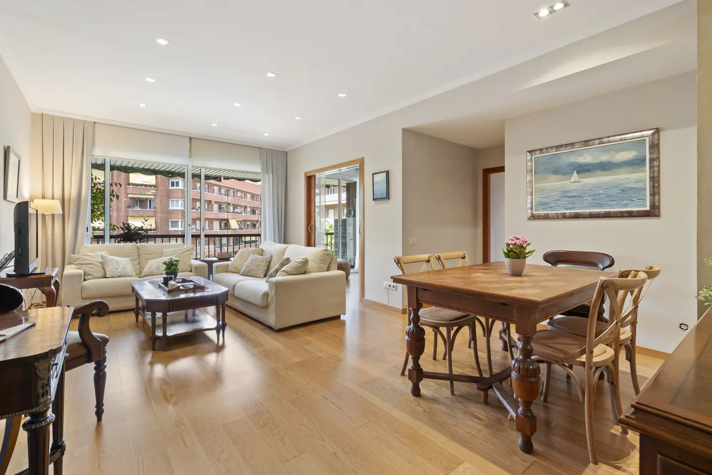 Bright living and dining area with hardwood floors. Cream sofas face a balcony. A wooden table with chairs sits near a painting of a sailboat.