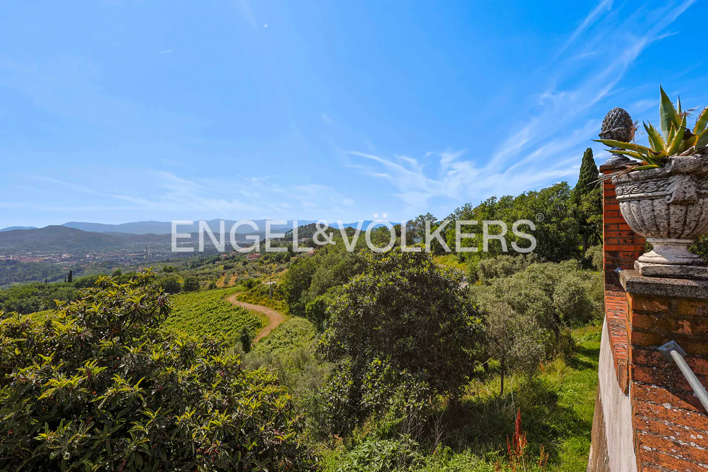 A stone planter with an agave plant overlooks a lush green valley, winding dirt path, and distant mountains under blue sky.