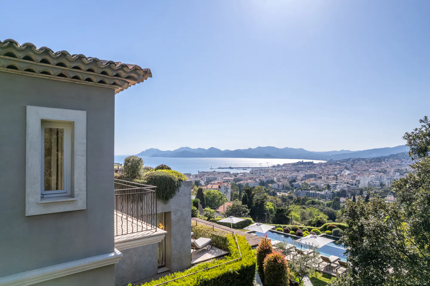 View of Cannes from a gray building with a balcony. A pool and lounge chairs are visible, with the city and mountains in the background.
