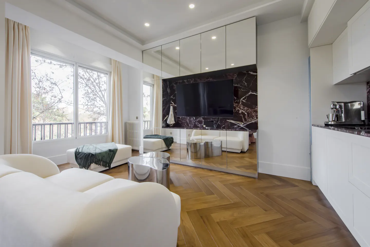 Bright living room with white sofa, herringbone wood floor, and mirrored wall with TV. Large windows with cream curtains.