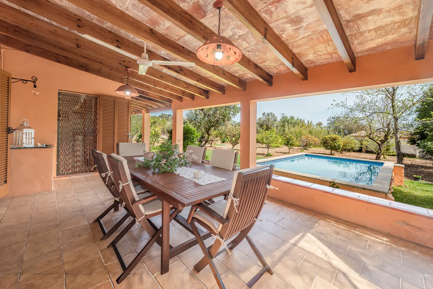 Covered patio with a wood table, chairs, and flowers. A pool and green trees are visible in the background.