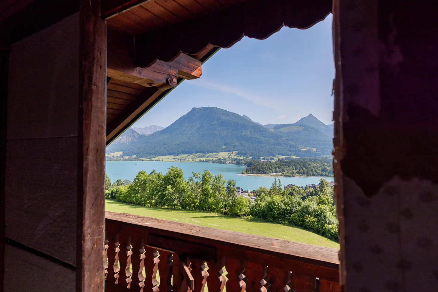 View from a wooden balcony overlooking a turquoise lake, green trees, and mountains under a blue sky.