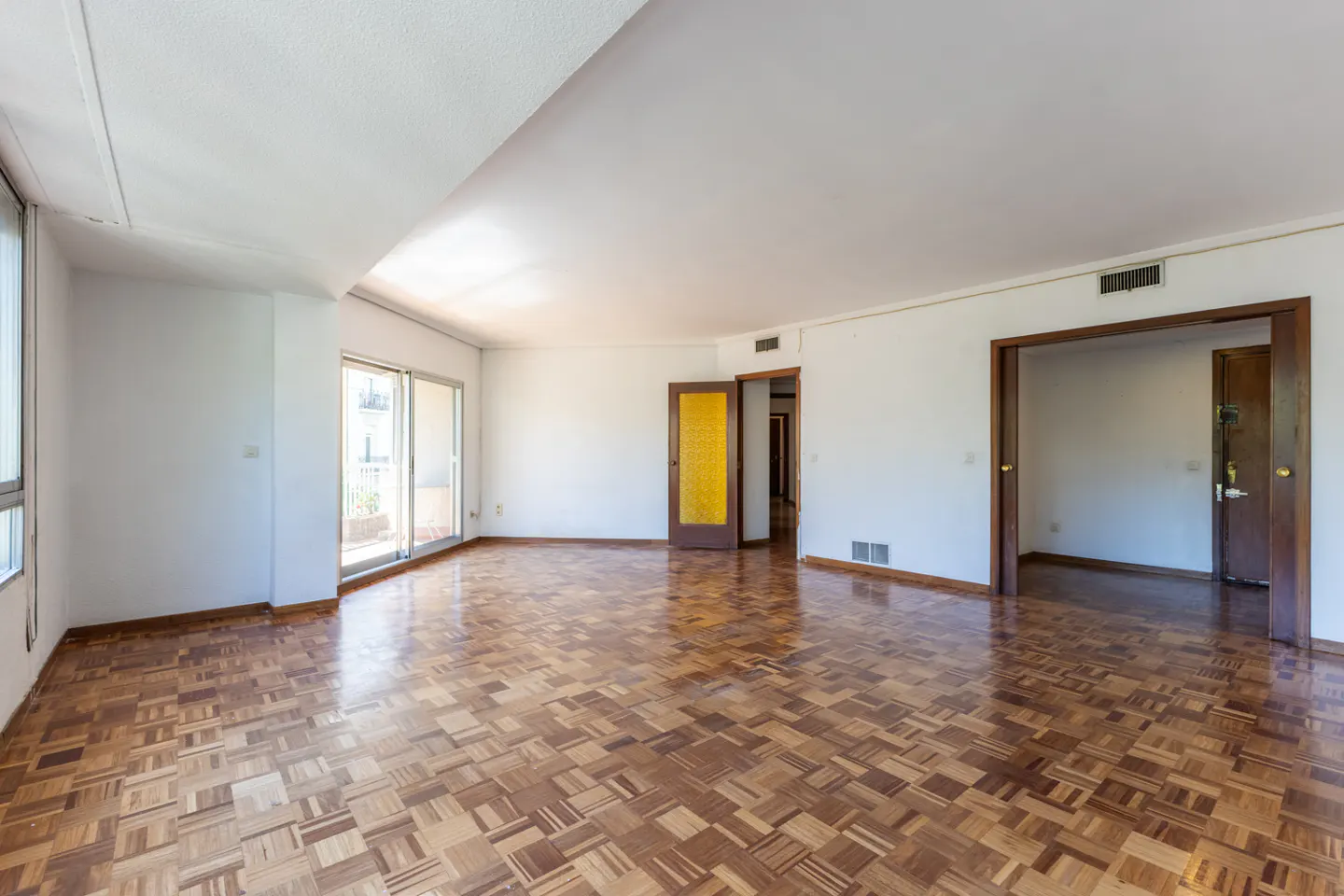 Empty living room with parquet floors, white walls, and doors leading to other rooms and a balcony.