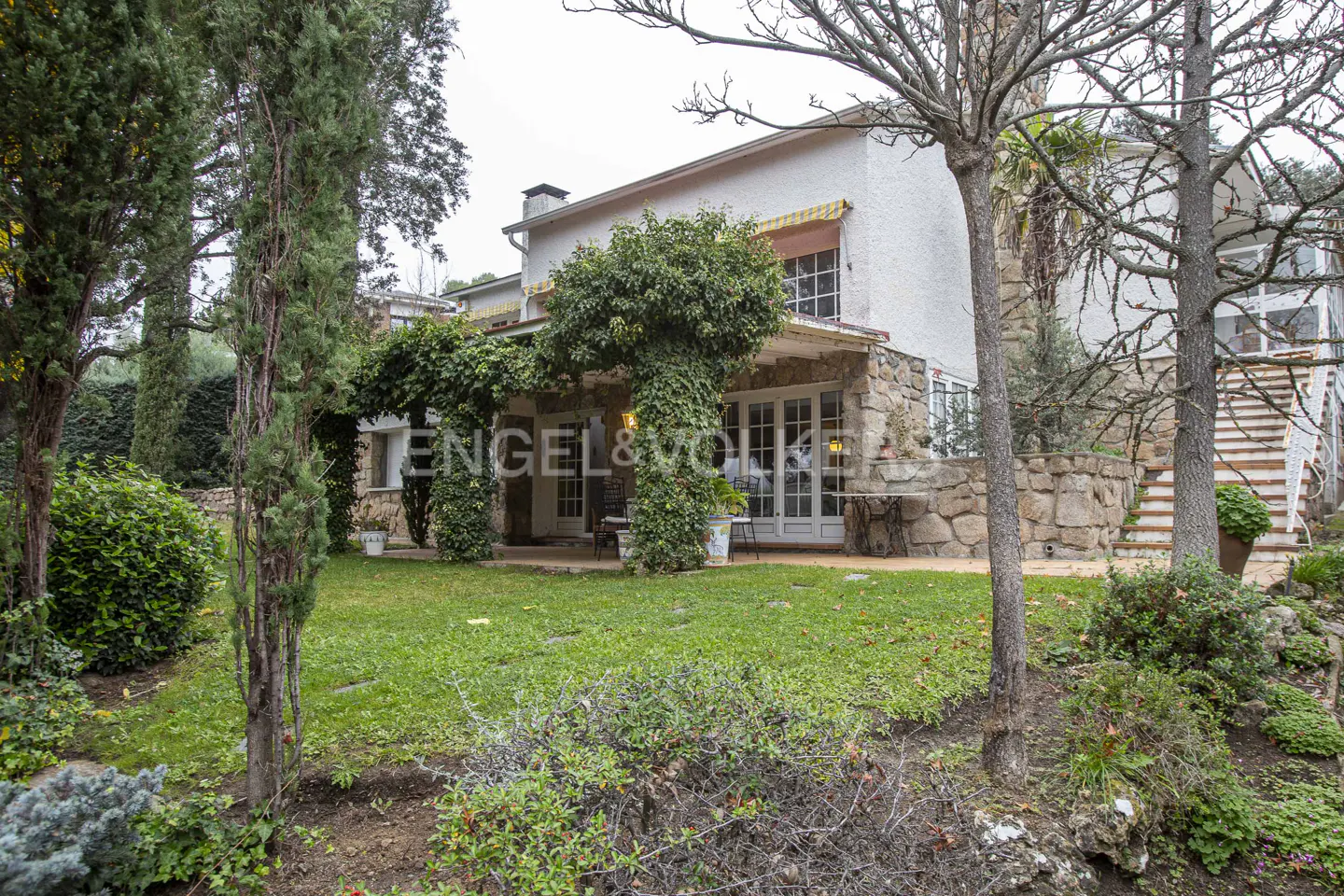 Exterior view of a two-story white house with stone accents and a green lawn. The porch is covered in ivy. The Engel & Völkers logo is visible.