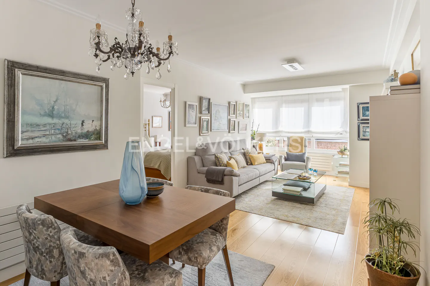 Bright living and dining room with a wooden table, crystal chandelier, grey sofa, and a gallery wall of framed art.