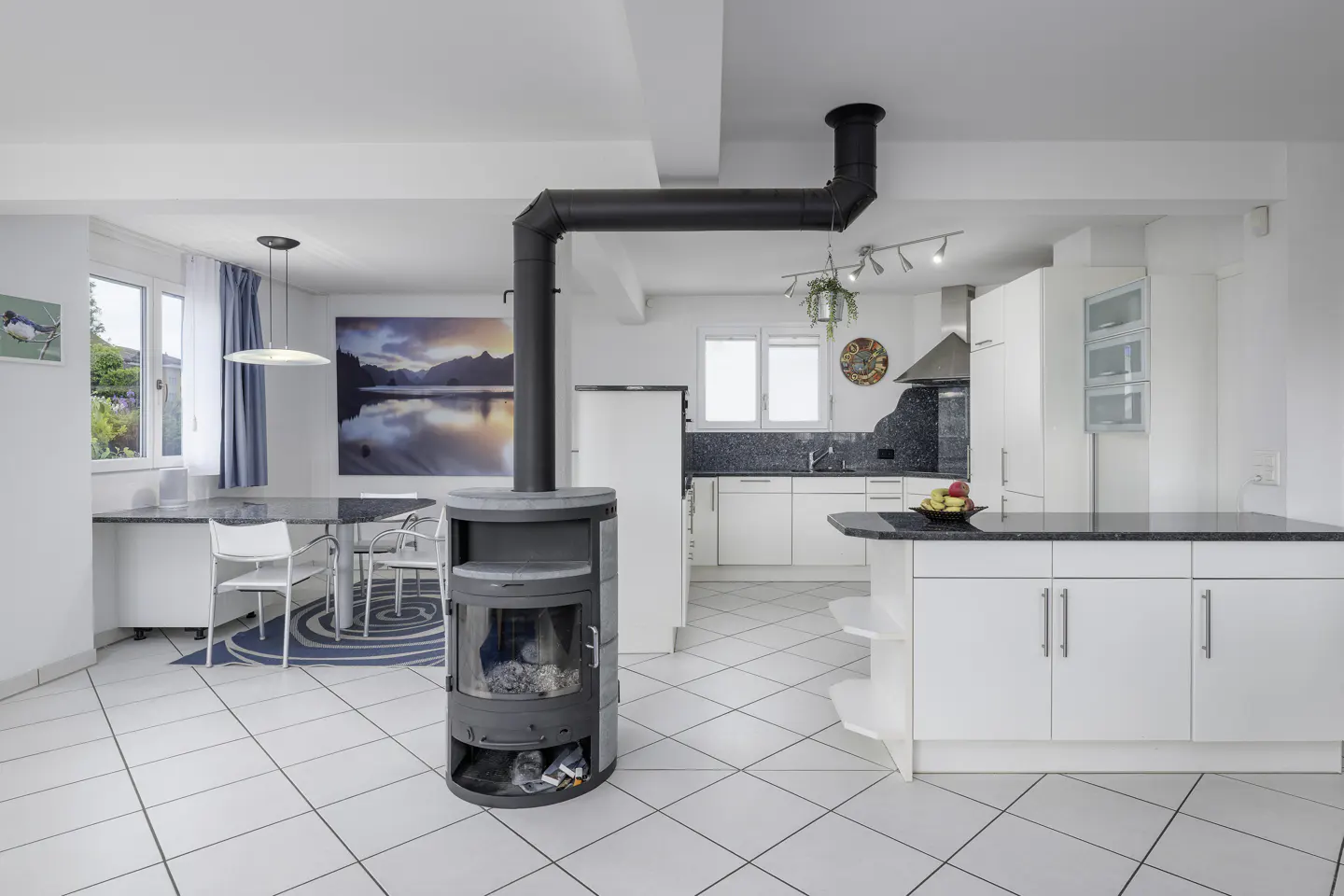 A bright, open-concept kitchen and dining area with white tile floors. A black wood-burning stove stands prominently in the foreground.