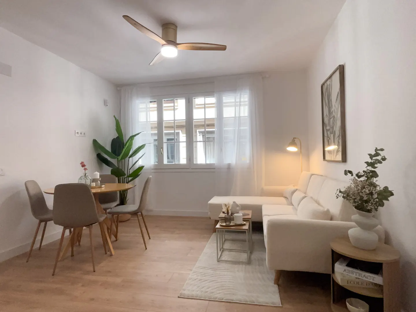 Bright living room with wood floors, white walls, and a ceiling fan. A round table with four chairs sits near a window. A white sofa and side tables are on a rug.