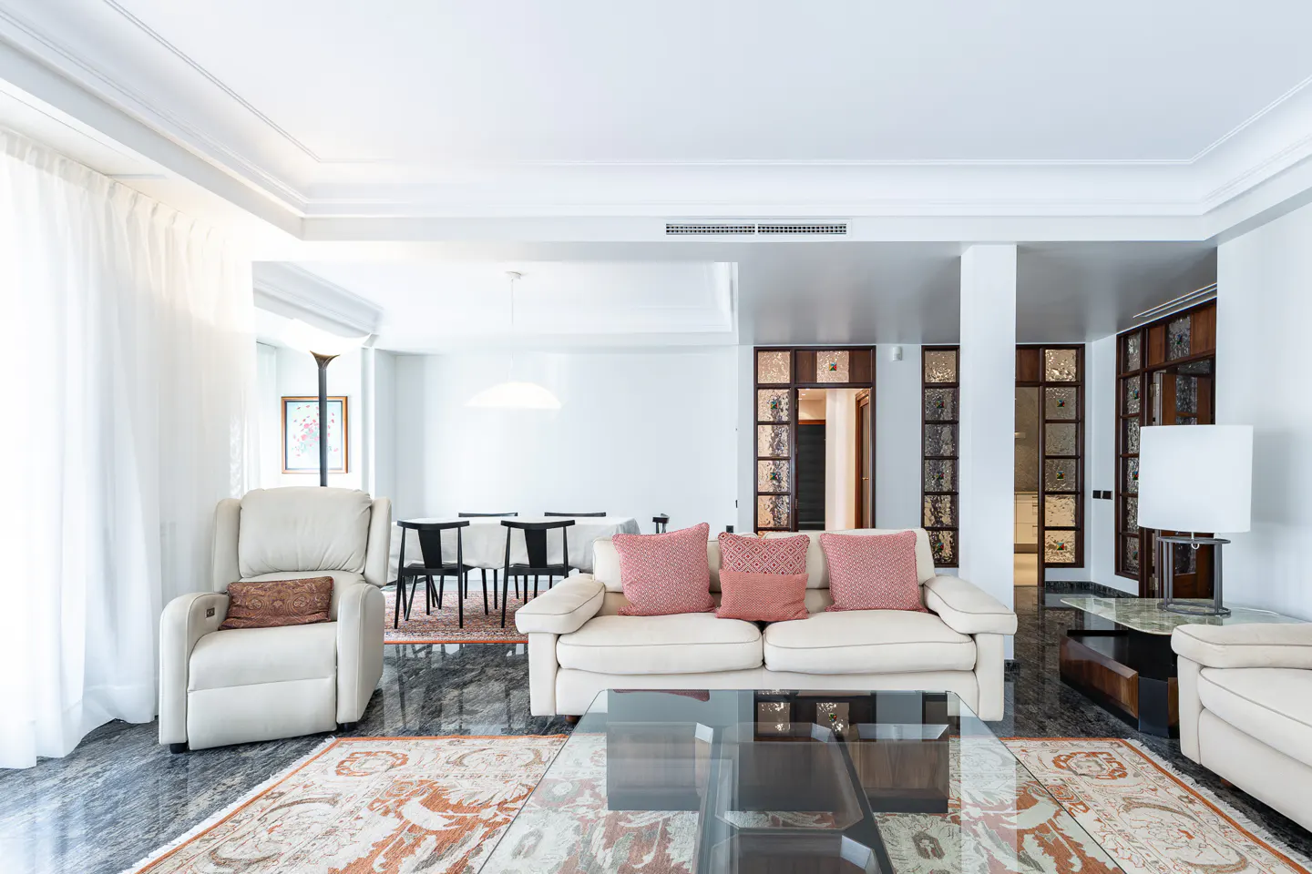 Bright living room with white walls, cream sofas, and pink pillows. A glass coffee table sits on a patterned rug over a marble floor. Dining area visible in the background.