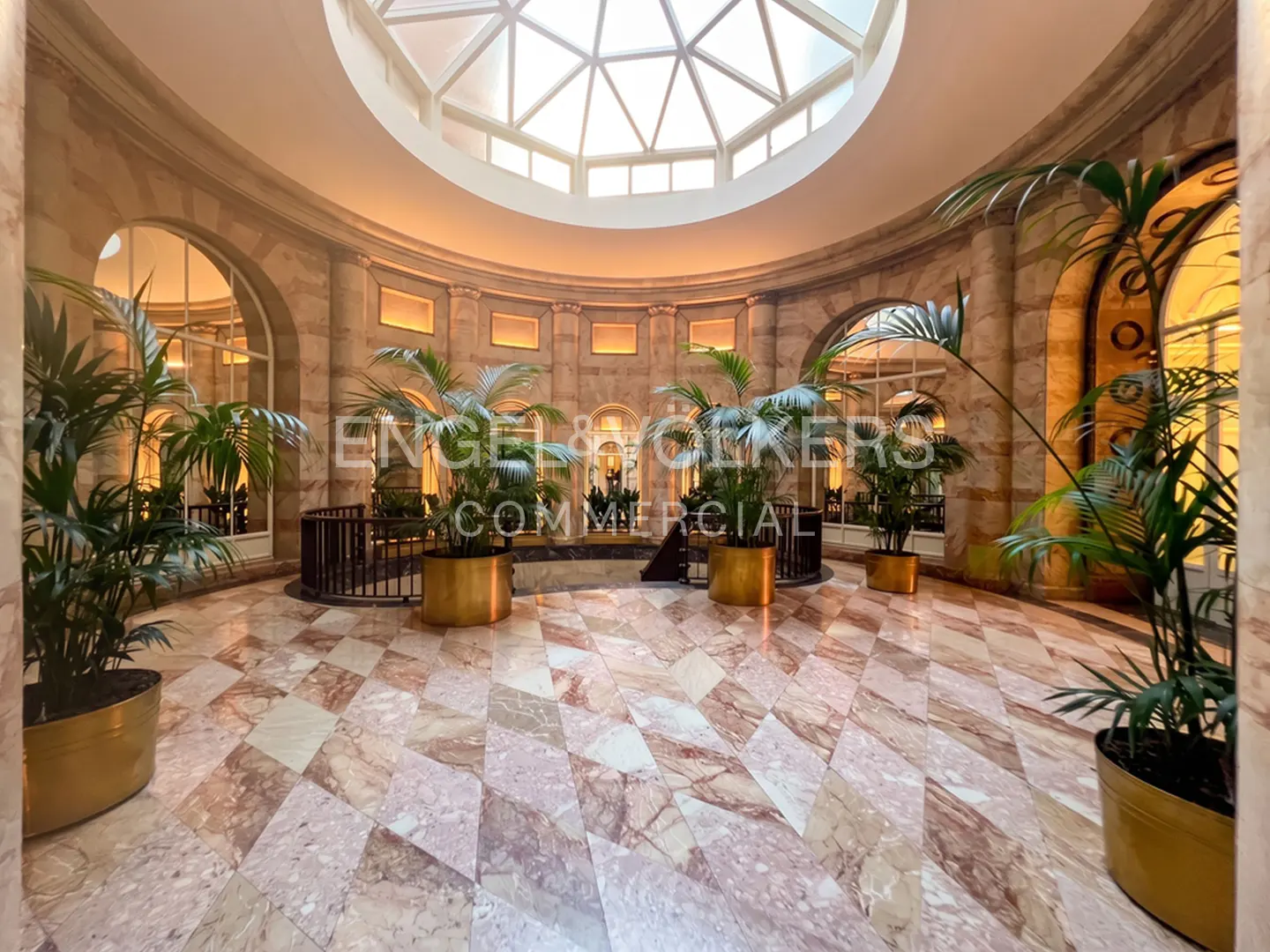 Interior view of a circular lobby with marble floors, potted plants, arched windows, and a domed skylight.