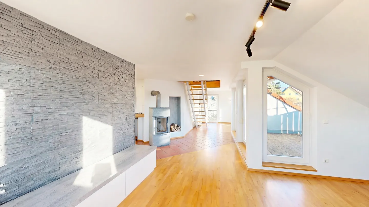 Bright, open living room with wood floors, stone accent wall, and fireplace. A ladder leads to an upper level. Windows offer a view of the outdoors.