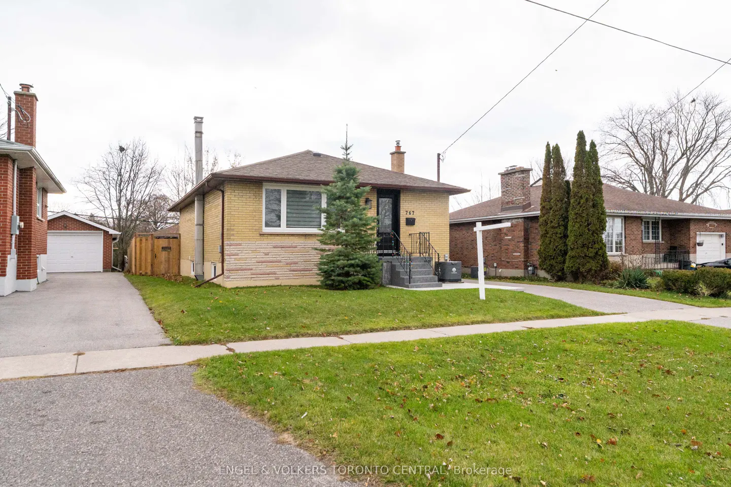 A yellow brick bungalow with a brown roof and a small evergreen tree in the front yard. A "For Sale" sign is visible.
