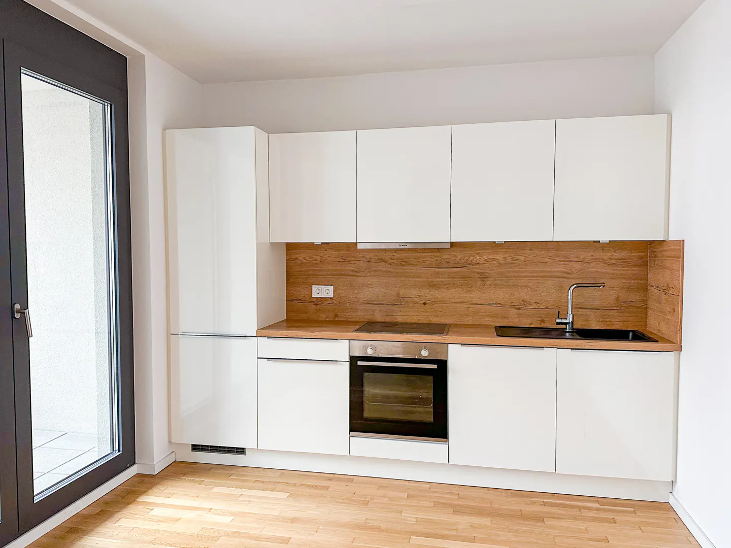 Bright kitchen with white cabinets, wood countertop and backsplash, stainless steel oven, and black sink. A large window is on the left.
