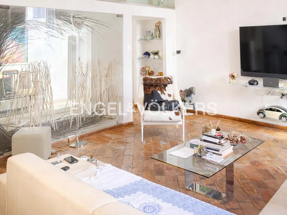 Living room with a glass wall, white walls, and a brown tiled floor. A glass coffee table sits in front of a white chair with a cowhide pillow. A TV is mounted on the wall.