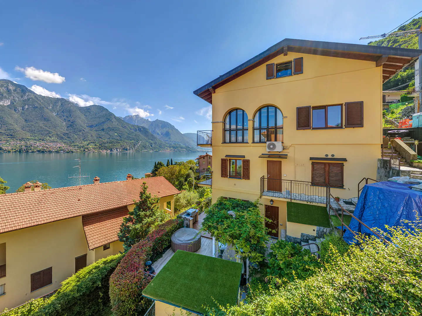 Exterior view of a yellow two-story house with brown shutters, overlooking a lake and mountains under a blue sky.
