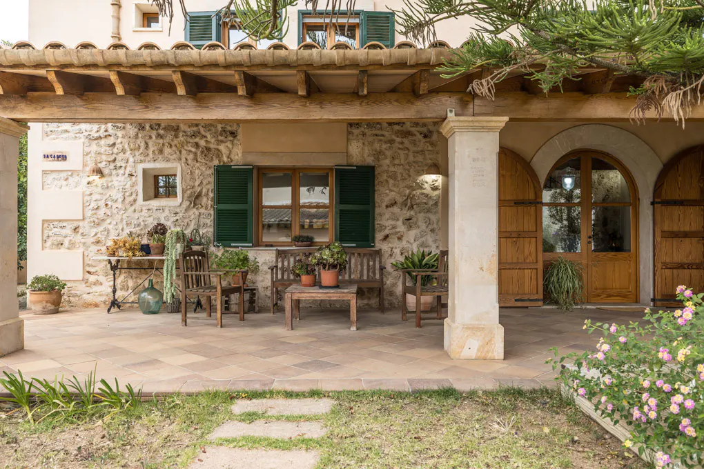 Stone house with a covered patio featuring wooden furniture, arched doors, and green shutters. Flowers and greenery add a touch of nature.