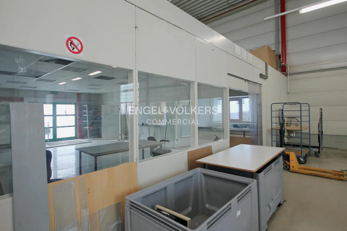 Interior view of a commercial space with a glass-enclosed office area, tables, storage bins, and a blue cart. "Engel & Völkers Commercial" logo on glass.