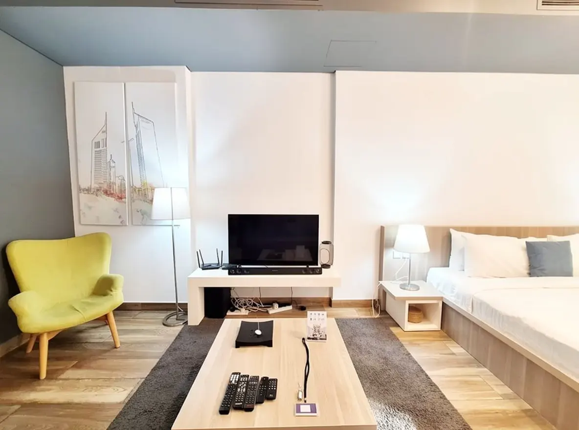 Living room with a yellow chair, TV, and bed. A light wood coffee table sits on a gray rug. Artwork hangs on the white walls.