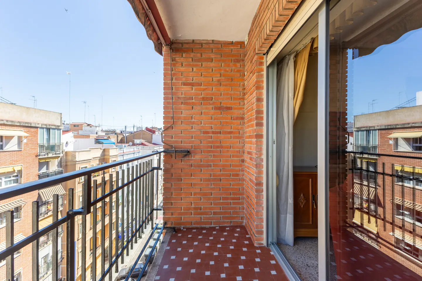 A balcony with a black railing, red brick wall, and red tile floor overlooks a city on a sunny day. Sliding glass doors lead inside.