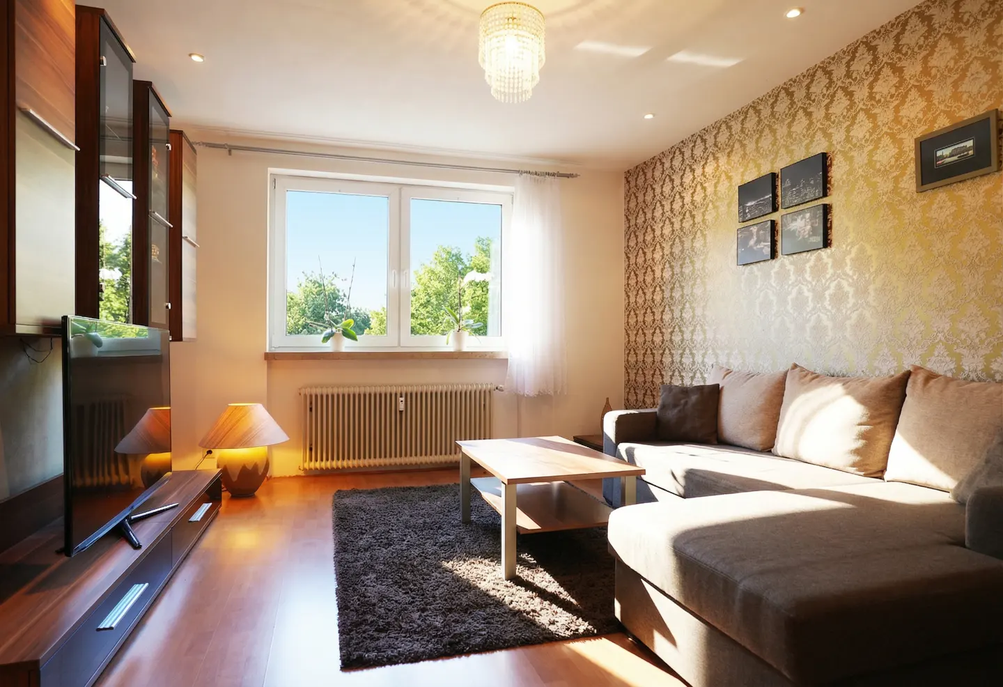 Living room with brown sectional sofa, wood floors, and patterned wallpaper. A window overlooks green trees.