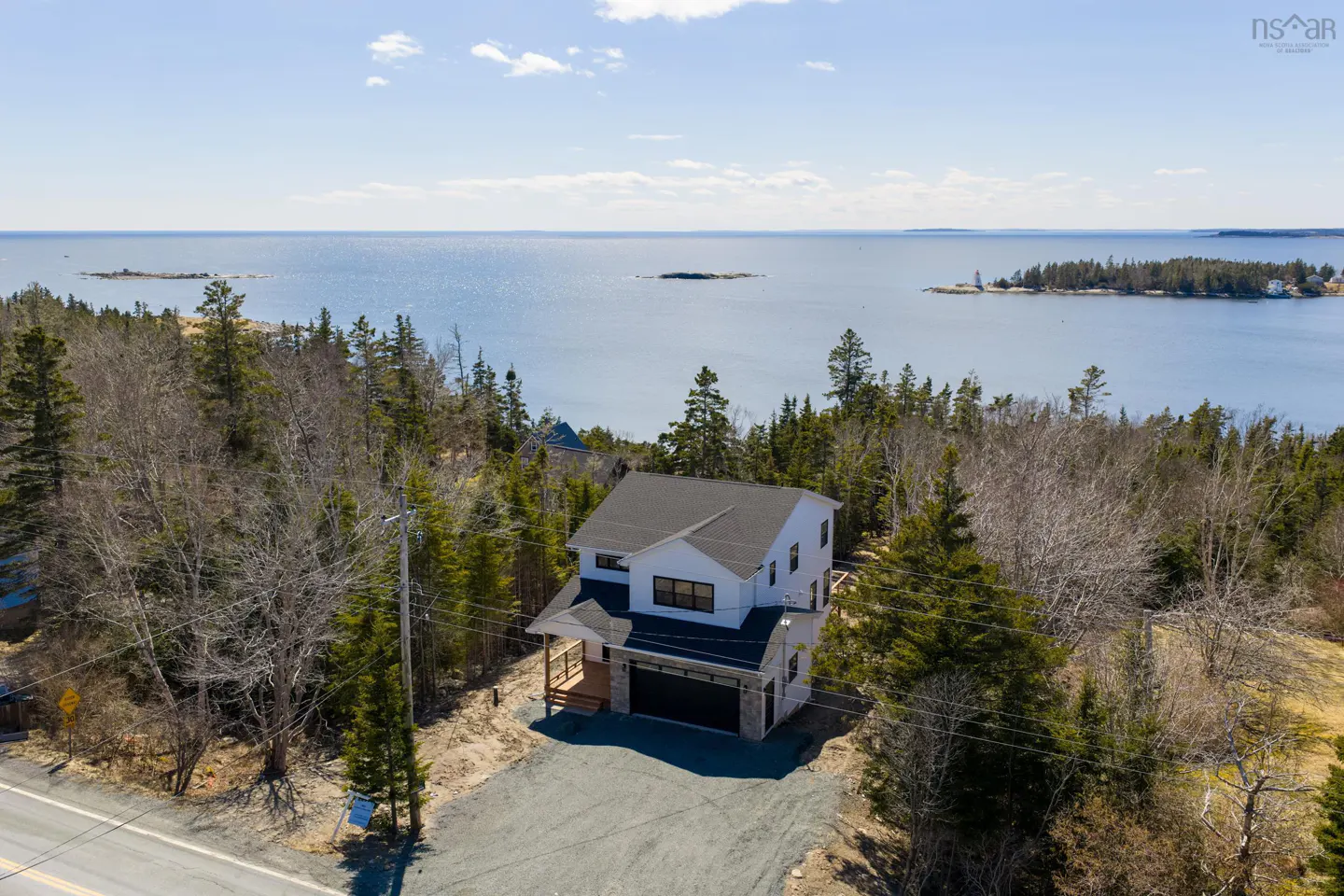 Aerial view of a modern white house with a black roof, surrounded by trees, overlooking a blue ocean with islands under a bright sky.