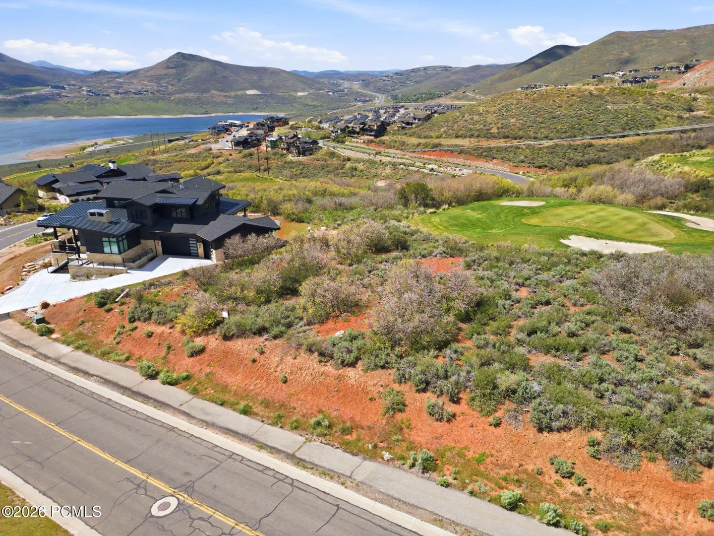A modern black house sits on a hill overlooking a lake and mountains under a blue sky.