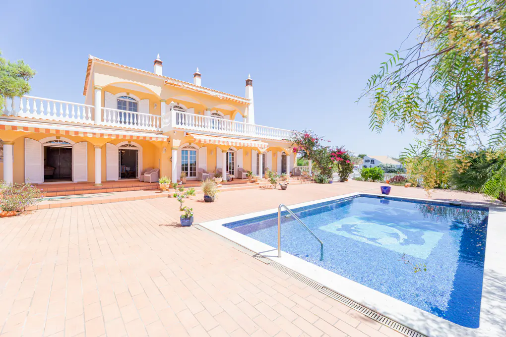 Two-story yellow villa with a white balcony, tiled patio, and a blue swimming pool under a clear blue sky.