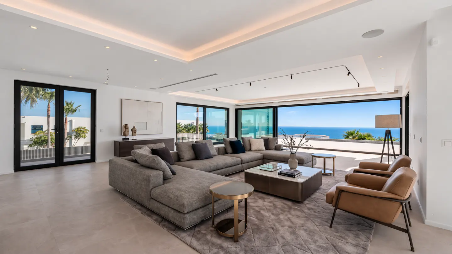 Bright living room with a gray sectional sofa, two tan chairs, and a view of the ocean through large windows.