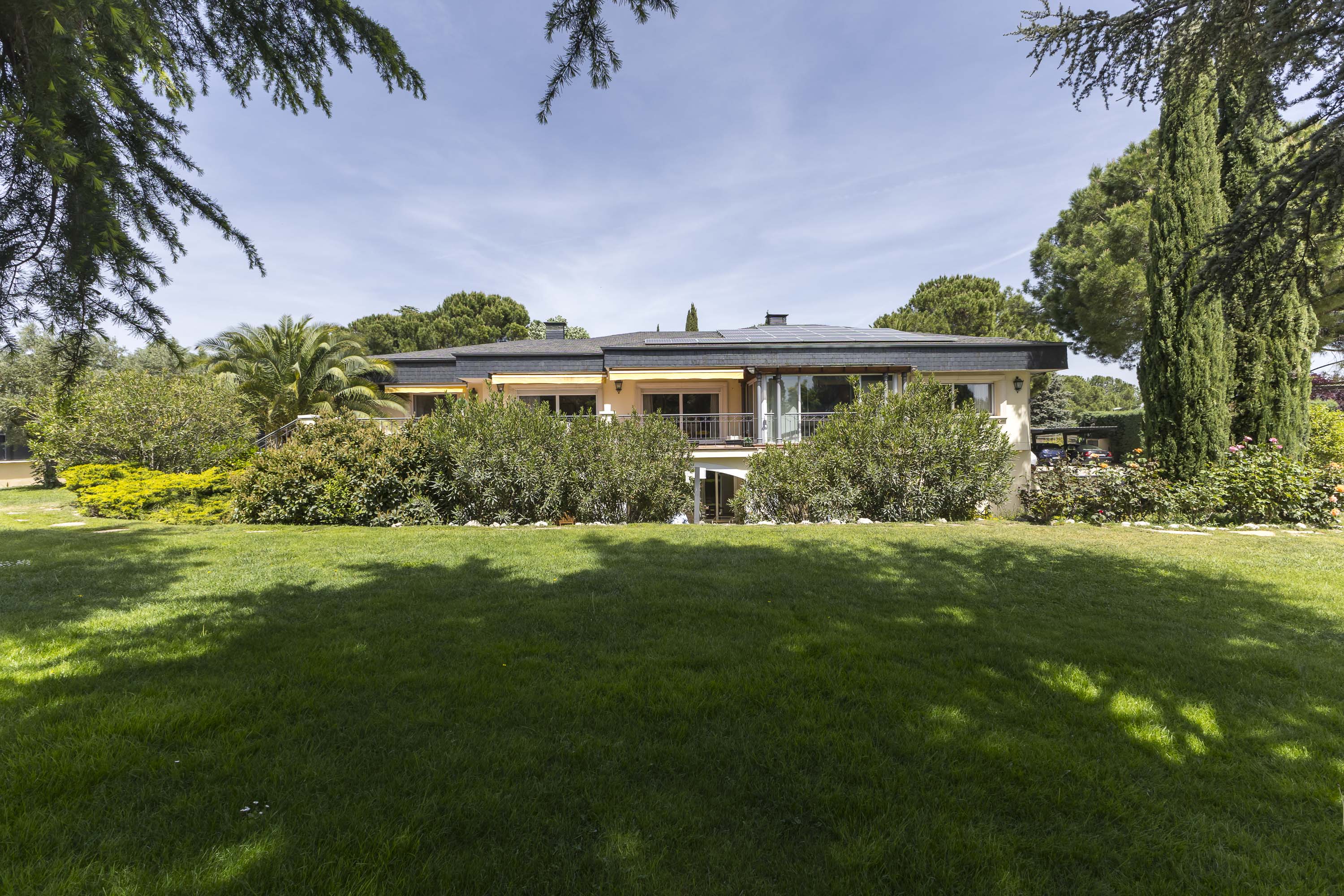 A tan house with a dark roof is surrounded by green grass and trees under a blue sky.