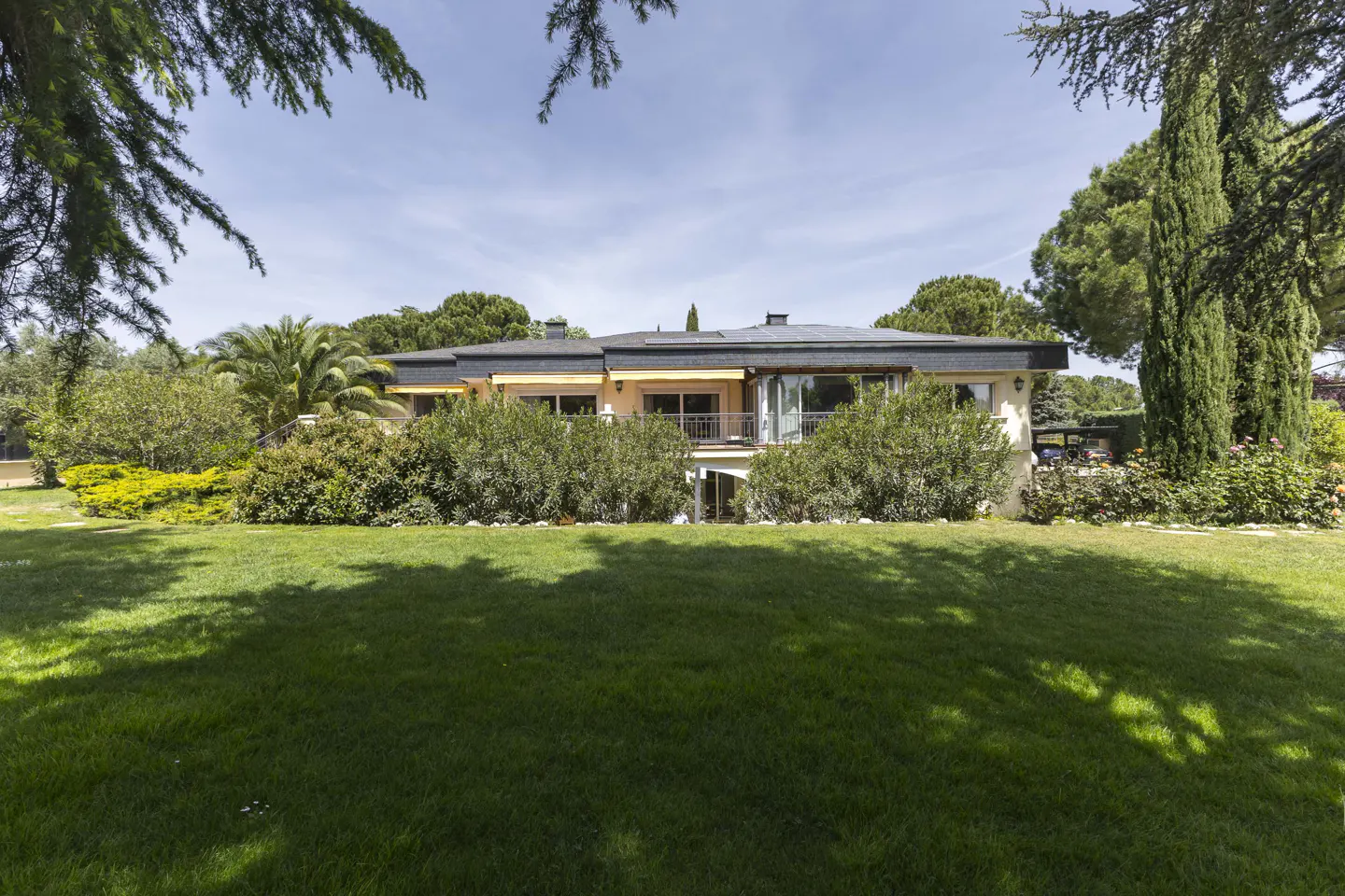 A tan house with a dark roof is surrounded by green grass and trees under a blue sky.