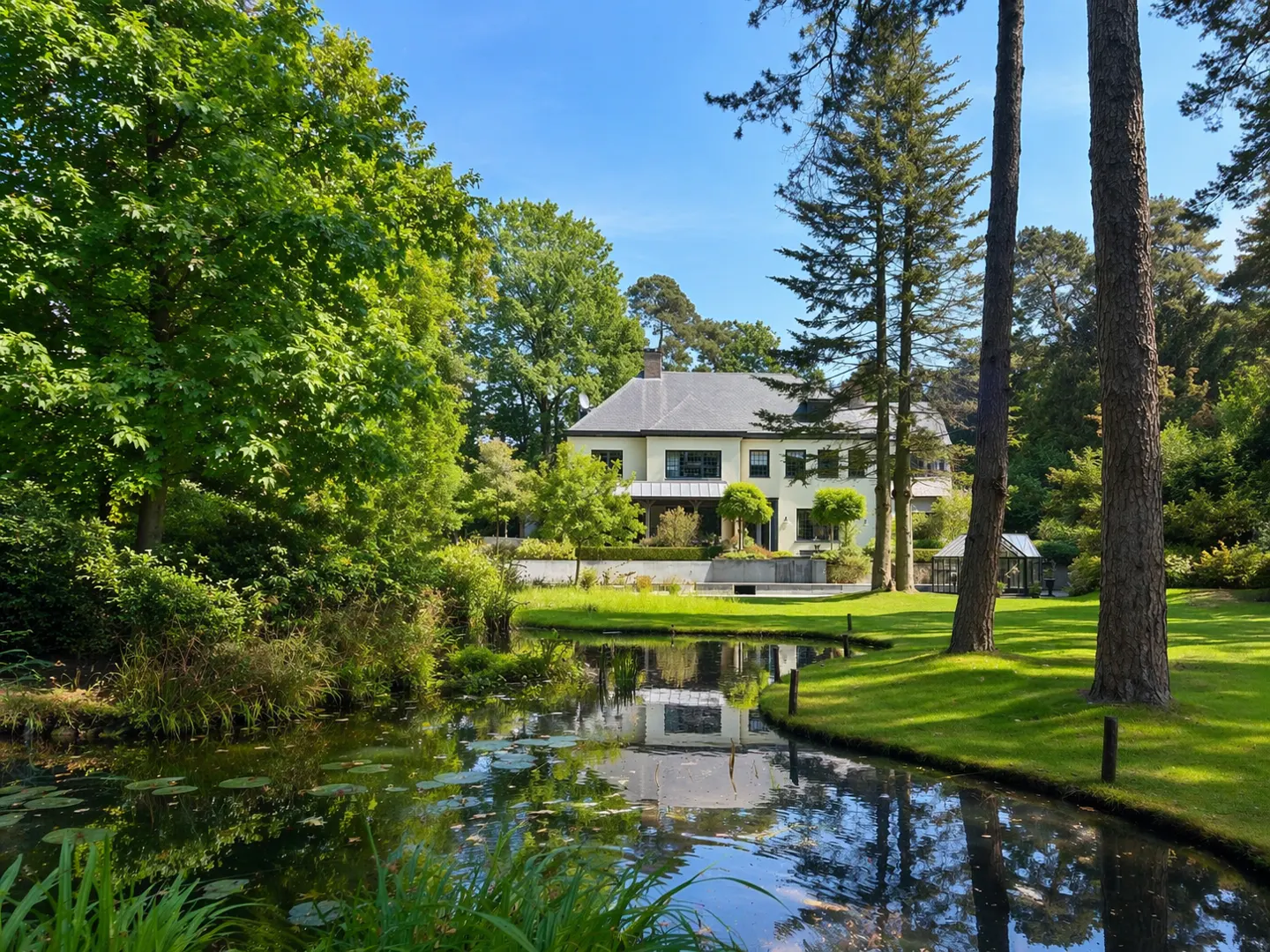 A large, light yellow house is surrounded by green trees and a pond reflecting the sky and house.