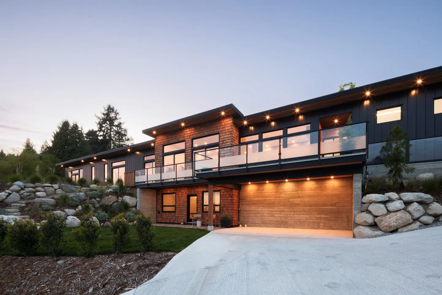 Modern two-story home with a concrete driveway, wood and dark siding, large windows, and a glass-railed balcony.