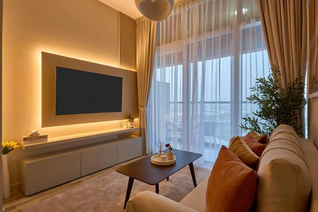 Living room with a TV, beige sofa with orange pillows, and a dark wood coffee table. Large windows with sheer curtains.