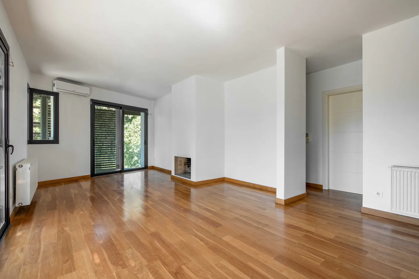 Bright, empty living room with hardwood floors, white walls, and black-framed windows and sliding doors. A fireplace is built into the wall.