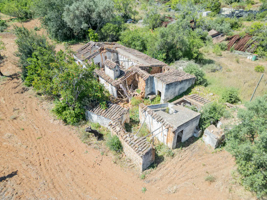 Aerial view of a dilapidated, white-walled building with a red tile roof, surrounded by green trees and brown, tilled soil.