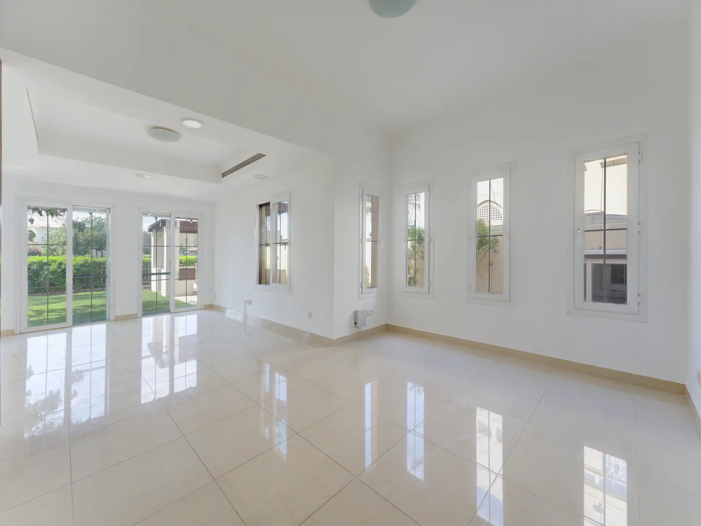 Bright, empty room with white walls, beige tile floor, and multiple windows. Sunlight reflects off the floor. A view of a green lawn is visible through the windows.